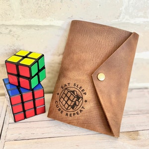 May include: A brown leather notebook with a flap closure and a gold button. The notebook has a black and white graphic of a Rubik's cube with the text "Eat Sleep Cube Repeat" around it.