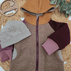 Könnte beinhalten: Eine braune, burgunderrote und rosafarbene Kapuzenjacke mit Reißverschluss. Die Jacke hat ein gestreiftes Muster an der Kapuze und am unteren Saum. Die Jacke wird mit einer gestreiften Mütze kombiniert.