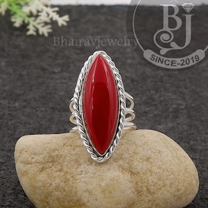 May include: A silver ring with a red oval-shaped gemstone set in a twisted silver band. The ring is on a light brown rock.
