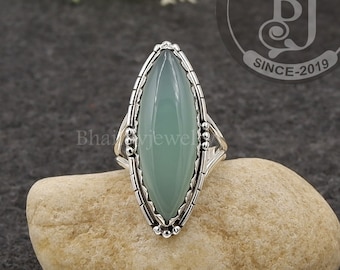 May include: A silver ring with a light green oval-shaped gemstone set in a detailed silver setting. The ring is on a rock.