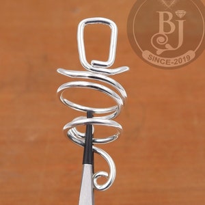 May include: Silver spiral ring with a square top. The ring is being held by a pair of pliers.