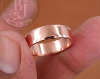 Puede incluir: Un anillo de oro rosa con una superficie lisa y pulida. El anillo se sostiene entre los dedos, mostrando su diseño de banda simple. La imagen incluye un logotipo que dice "BJ SINCE-2019".