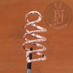 May include: A rose gold spiral ring with an engraved floral pattern. The ring wraps around the finger in a unique design. The ring is held by a black tool against a brown background. The logo "BJ SINCE-2019" is in the background.
