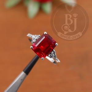 Può includere: Un anello d'argento con una grande gemma rossa rettangolare come elemento centrale. L'anello è impreziosito da gemme più piccole e trasparenti ai lati della pietra principale. L'anello è tenuto da una pinzetta.