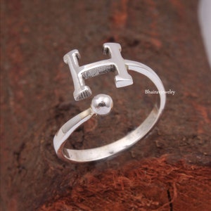May include: Silver adjustable ring with a letter H charm. The ring is open and can be adjusted to fit different finger sizes.
