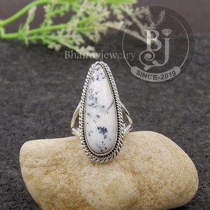 May include: A silver ring with a large, teardrop-shaped white stone speckled with black. The stone is framed by a twisted rope-like border and set on a silver band. The ring is displayed on a light brown rock.