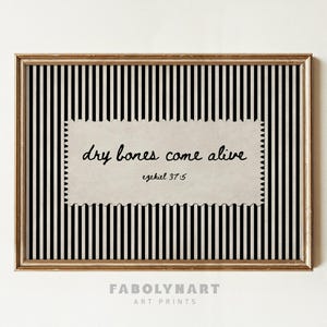 May include: A framed wall art print featuring a black and white vertical striped background. Centered is a rectangular beige paper cutout with scalloped edges displaying the black handwritten text: dry bones come alive ezekiel 37:5.