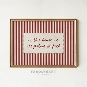 May include: A framed art print featuring a red and white vertical striped background. Centered is a cream-colored, scalloped-edge rectangular label with the dark red text: in this house we are festive as fuck. The frame is a metallic gold color.