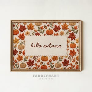 May include: A rectangular framed art print featuring a central cream-colored panel with the text "hello autumn" in brown script. The border is decorated with an embroidered-style pattern of orange pumpkins, red and yellow maple leaves, and acorns.