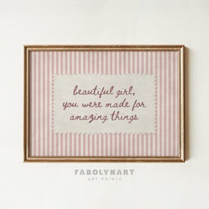 May include: A rectangular wall art print in a gold frame featuring a light pink and white vertical striped background. Centered is a cream-colored scalloped-edge rectangle with the dark pink text: beautiful girl, you were made for amazing things.