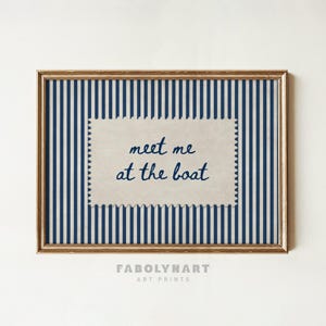 May include: A rectangular wall art print in a gold frame featuring a vertical navy blue and white striped background. Centered is a beige, postage stamp-shaped paper cutout with the dark blue handwritten text meet me at the boat.