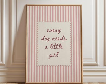 Puede incluir: Una lámina artística enmarcada con un fondo de rayas verticales en rosa claro y blanco. En el centro, un panel rectangular de color crema con bordes festoneados contiene el texto en rojo oscuro: every dog needs a little girl. El marco es dorado.