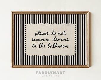 May include: A framed wall art print featuring a vertical black and white striped background. Centered is a cream-colored, postage stamp-shaped paper with the black text: please do not summon demons in the bathroom. The frame is a metallic gold color.