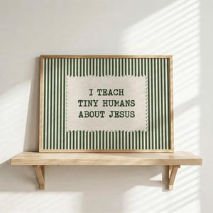 May include: A rectangular framed print featuring a dark green and white vertical striped background. Centered is a cream-colored rectangular label with scalloped edges containing the black text: I TEACH TINY HUMANS ABOUT JESUS.