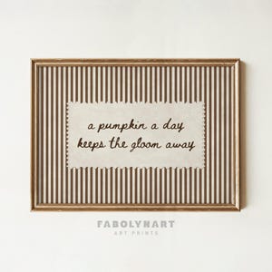 May include: A framed wall art print featuring a cream-colored rectangular paper with scalloped edges centered on a brown and white vertical striped background. The text reads: a pumpkin a day keeps the gloom away in a dark brown cursive font.