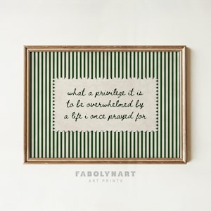 May include: A rectangular wall art print in a metallic gold frame featuring a dark green and white vertical striped background. Centered is a cream-colored paper cutout with scalloped edges displaying the black text: what a privilege it is to be overwhelmed by a life i once prayed for.