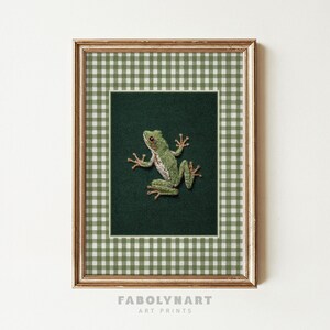 May include: Framed art print featuring a detailed embroidered frog on a dark green background. The frog is light green with brown accents. The frame has a gold finish and a green and white gingham pattern.