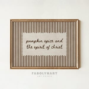 May include: A rectangular wall art print in a metallic gold frame featuring a brown and cream vertical striped background. A central cream-colored rectangular paper cutout with scalloped edges displays the text pumpkin spice and the spirit of christ.