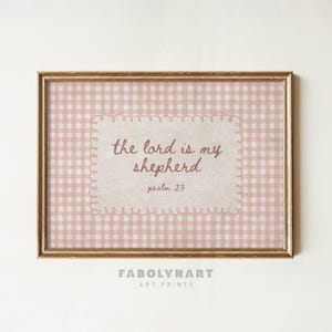 May include: A rectangular wall art print in a gold frame featuring a light pink and white gingham pattern background. Centered is a cream-colored scalloped label with the brown text: the lord is my shepherd, psalm 23. The design is clean and simple.