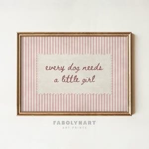 May include: A rectangular wall art print in a gold frame featuring a light pink and white vertical striped background. Centered is a cream-colored rectangular paper cutout with scalloped edges displaying the text every dog needs a little girl.
