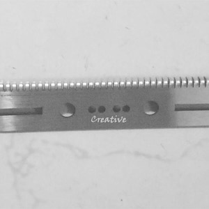 Puede incluir: Una herramienta de metal plateado con una serie de pequeños dientes a lo largo de un borde. La herramienta tiene forma rectangular con varios agujeros y ranuras. La palabra "Creative" está grabada en el metal. Diseñada para un oficio o uso industrial específico.