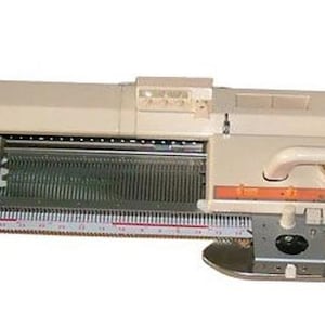 May include: A cream-colored knitting machine with a long, rectangular body. It features a handle, a ruler, and a perforated section. The machine has a white handle and a dial. The text "CREATIVECOUNTER" is visible.