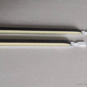 Puede incluir: Dos objetos alargados y paralelos con una franja negra y una franja amarillo claro, y un extremo rectangular blanco. Los objetos están sobre una superficie gris claro. Los objetos parecen ser parte de un artículo más grande.