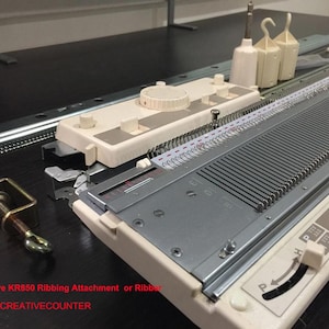 May include: A beige and silver Creative KR850 ribbing attachment for a knitting machine, featuring a metal needle bed, a control dial, and two white plastic weights. The unit includes a metal clamp and is labeled with the text Creative KR850.