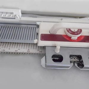 May include: Close-up of a white and red knitting machine. The machine has a handle, a gauge, and a series of needles. The machine is designed for creating knitted fabric. The machine is on a light gray surface.