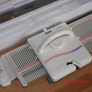 May include: A white knitting machine, model LK150, with a handle and a dial. The machine has pink and purple accents and is partially open, revealing the needles. The machine is in its original packaging.