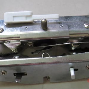 May include: A silver-colored sewing machine attachment with a white plastic lever and a small white button. The attachment has a rectangular shape with various metal components and screws. The text "CREATIVECOUNTER" is visible in red.