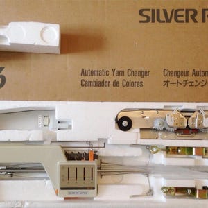 May include: A Silver Reed YC 6 automatic yarn changer machine, packaged in a cardboard box with foam inserts. The machine is beige and silver, with various mechanical components visible. The box also includes text in multiple languages.