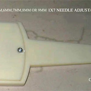 May include: An off-white plastic 1x7 needle adjustable transfer tool with seven metal needles on one end and a single needle on the other. The tool features the text 6.5MM, 6MM, 7MM, 8MM OR 9MM 1X7 NEEDLE ADJUSTABLE TRANSFER TOOL and CREATIONGROUPS.