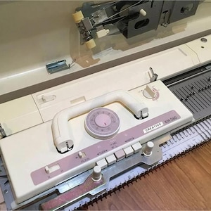 Può includere: Una macchina per maglieria Creative KH-260 vintage in crema e rosa. La macchina presenta una maniglia bianca, un quadrante con numeri e diversi pulsanti. Il testo "CREATIVE KH-260" è visibile. La macchina è su una superficie di legno.
