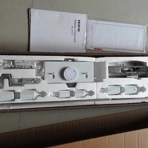 May include: A new, white sewing machine attachment kit, neatly arranged in a cardboard box with protective foam inserts. The kit includes various metal and plastic components, a dial, and a manual. The box is open, revealing the contents.