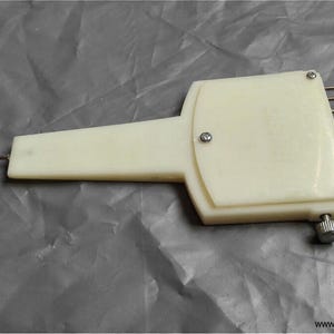 Puede incluir: Una herramienta de transferencia para máquina de tejer de plástico color crema con un mango ancho, una aguja de metal en la parte frontal y ocho agujas de metal paralelas con ojales en el lado opuesto, aseguradas por tres tornillos pequeños.