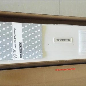 以下が含まれることがあります： 段ボール箱に入った新品のシルバーリード編み機、モデルSK155。保護フォーム付き。取扱説明書と書類が見えます。箱には「CREATIVECOUNTER」と記載されています。
