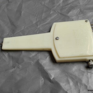 Puede incluir: Una herramienta de tejido vintage de color blanquecino con un mango largo y una serie de agujas de metal. La herramienta tiene un cuerpo rectangular con una perilla y está asegurada con tornillos pequeños. El fondo es una superficie gris arrugada.