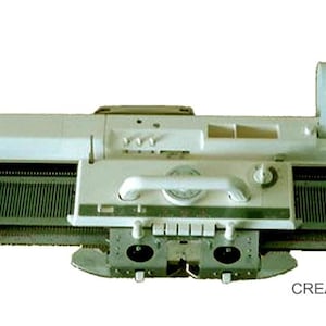 Puede incluir: Una máquina de tejer blanca y gris con una larga cama de agujas de metal, un carro deslizante con asa y un mecanismo lector de tarjetas perforadas montado en el lado derecho. El dispositivo incluye varios mandos y palancas de control.