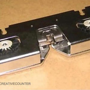 Puede incluir: Un dispositivo metálico plateado y negro con dos componentes circulares y detalles rojos. El dispositivo rectangular tiene una sección articulada en el centro. El texto "CREATIVECOUNTER" es visible en la esquina inferior izquierda.