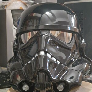 Può includere: Un casco nero lucido con visiera riflettente, che ricorda un design da stormtrooper. Il casco presenta dettagliate modanature, tra cui una prominente griglia per la bocca e accenti laterali. È esposto su una superficie di legno.