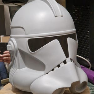 Könnte beinhalten: Ein weißer Plastik-Klonkrieger-Helm mit schwarzem Visier. Der Helm steht auf einer Holzoberfläche mit zwei kleinen weißen Plastikteilen.