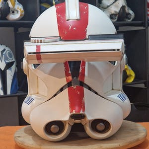 May include: A white and red helmet with a visor, featuring a weathered appearance. The helmet has a central red stripe and blue accents. It is displayed on a wooden stand, with other helmets visible in the background.