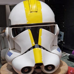 May include: A white and yellow Star Wars clone trooper helmet with blue markings. The helmet has a visor and a detailed design.