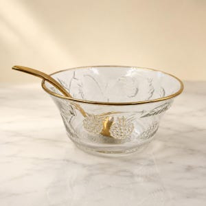 May include: A clear glass bowl featuring an embossed pineapple pattern and a gold-toned rim, accompanied by a matching gold-toned spoon resting inside. The bowl has a wide, circular opening and a smooth, transparent base with textured fruit details.