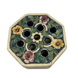 May include: An octagonal ceramic flower frog with a cream-colored border. The blue interior features floral and leaf designs in yellow, red, and green. The frog has multiple holes for arranging flowers.