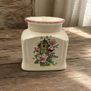 May include: A cream-colored ceramic jar with a wide opening and a pink speckled rim. The front features a colorful illustration of a birdhouse, a small brown bird, pink magnolia flowers, green leaves, and small blue berries on a vine.