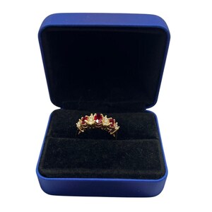 May include: A gold ring with a central oval ruby and smaller rubies and diamonds. The ring is displayed in a blue velvet-lined jewelry box with a black interior.