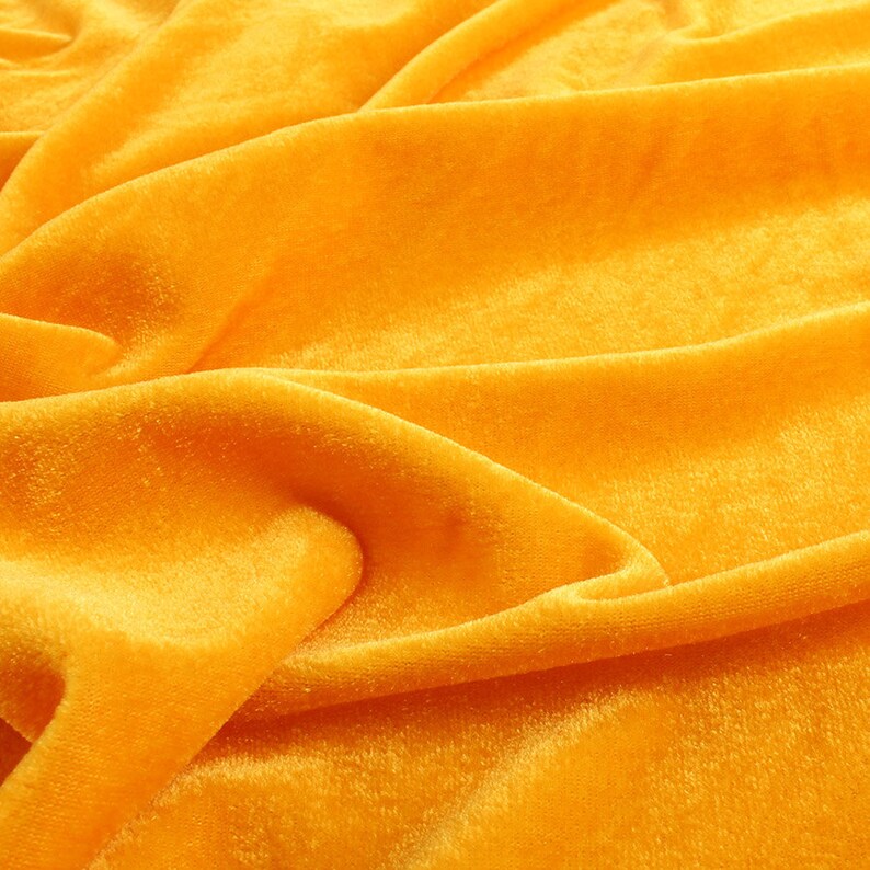 Op de afbeelding: Een close-up van een zachte, gele fluwelen stof met een glad, getextureerd oppervlak. De stof is gedrapeerd en gevouwen, wat een luxe en elegante uitstraling cre&euml;ert.