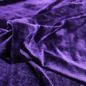 Op de afbeelding: Een close-up van een dieppaarse fluwelen stof met een zachte, pluche textuur. De stof is gedrapeerd en gevouwen, wat een luxe en elegante uitstraling cre&euml;ert.